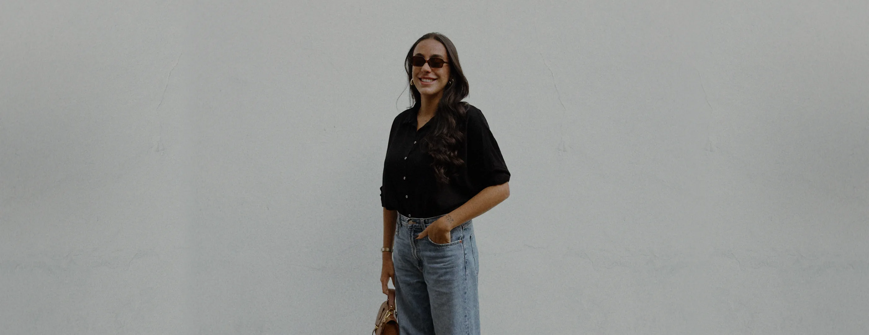 Anna Gargalak encostada em uma parede cinza, de camisa preta e calça jeans